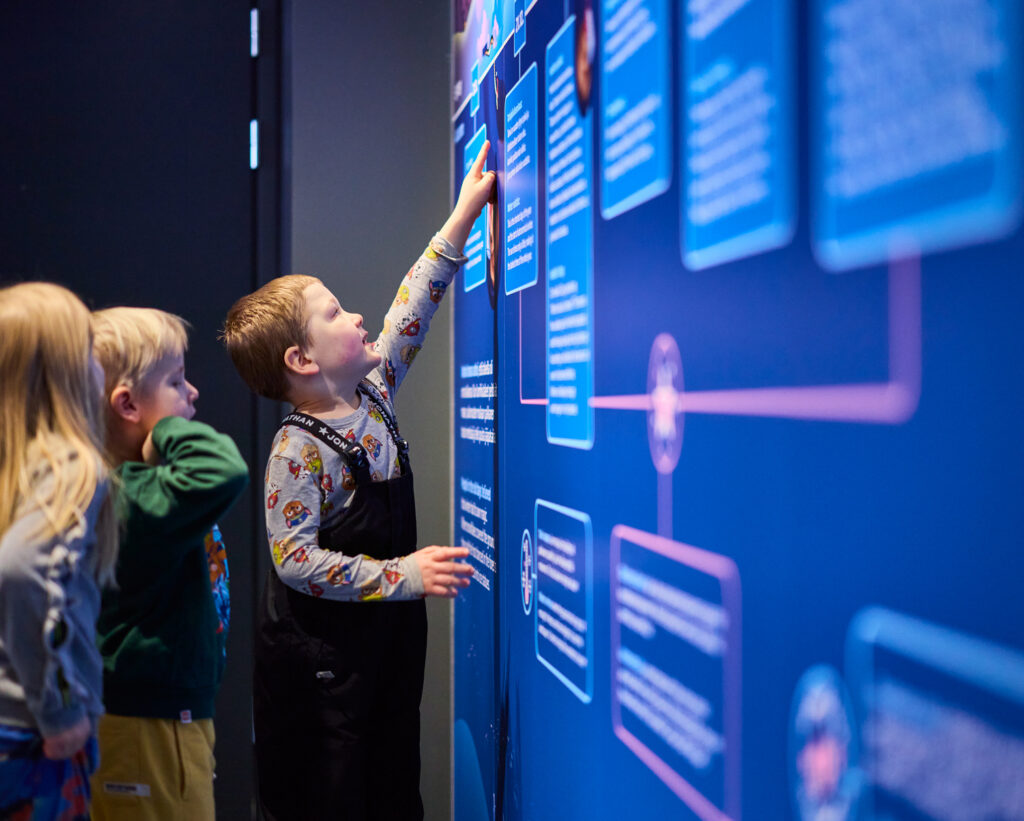 Lapsia tutustumassa Lumi-näyttelyyn. Poika osoittaa sormellaan näyttelyssä olevaa talven aikajanaa, kohdassa jossa kyykäärme painuu maan alle unimyssy päässään.