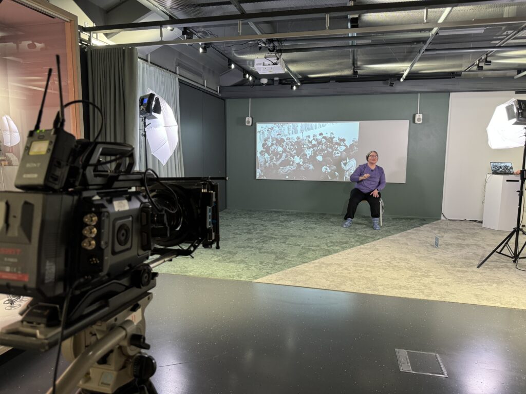 Värikuva kamerasta, joka kuvaa liikuntaharjoituksen ohjaajaa sisätiloissa. Ohjaajan taustalla näkyy seinälle heijastettu arkistokuva.